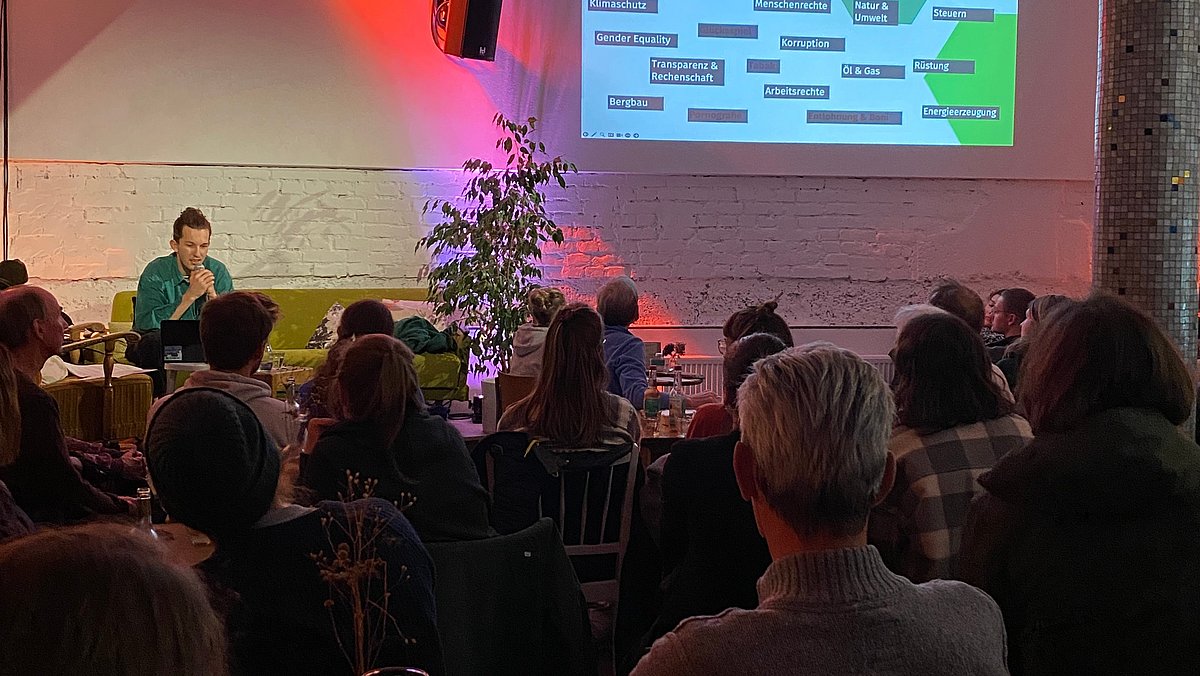 Referent Tobias referiert vor den Zuhörerinnen. Im Hintergrund eine Projektion zum Thema: Was macht eine konventionelle Bank nicht mit meinem Geld? 