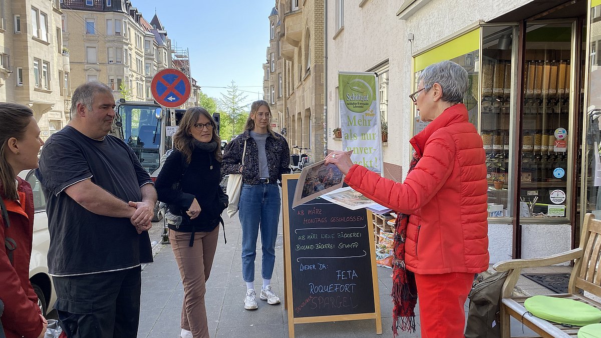 Menschen hören einer Referentin zu, sie stehen vor einem Laden im Rahmen einer Stadtführung