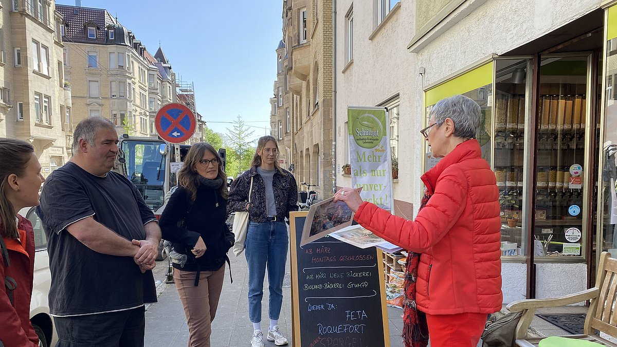 Menschen hören einer Referentin zu, sie stehen vor einem Laden im Rahmen einer Stadtführung