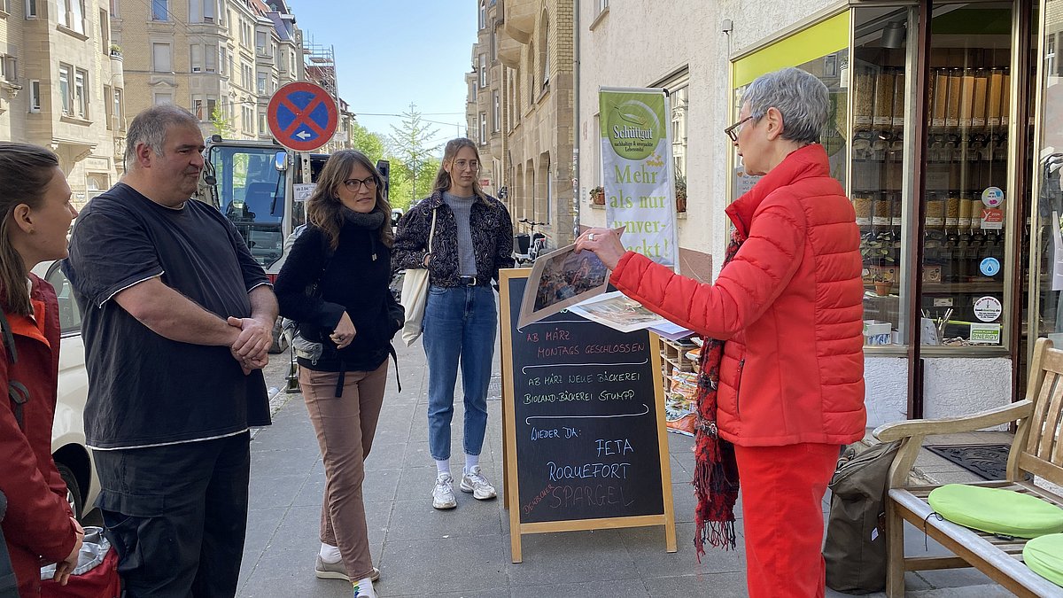 Menschen hören einer Referentin zu, sie stehen vor einem Laden im Rahmen einer Stadtführung