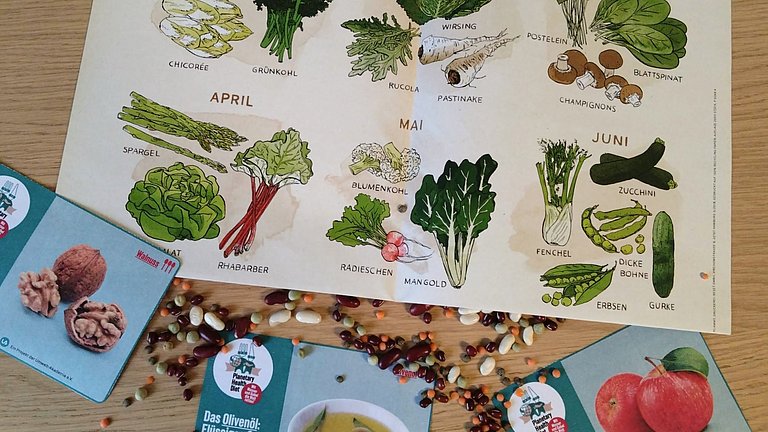 Ein Saisonkalender liegt auf einem Tisch, umgeben von Bohnen und verschiedenen anderen Samen.