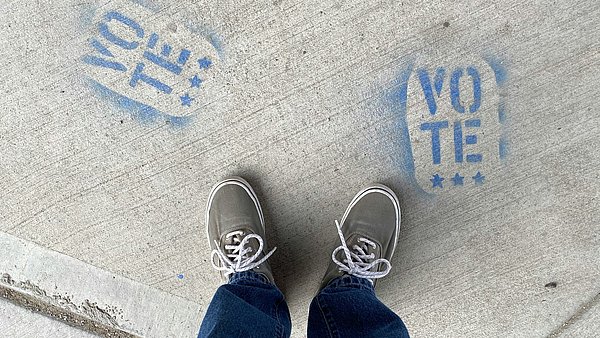 Zwei Beine einer Person stehen auf Asphalt, vor ihnen Bodengraffitis mit der Aufschrift "Vote"