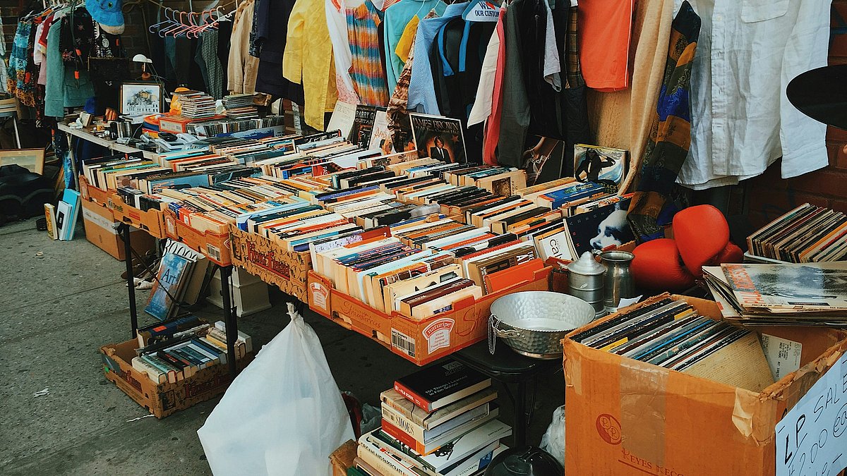 Flohmarktstand mit einer großen Auswahl an gebrauchten Büchern, Schallplatten, Kleidung und Haushaltsgegenständen, die auf Tischen und Kleiderstangen zum Tauschen oder Weitergeben präsentiert werden.