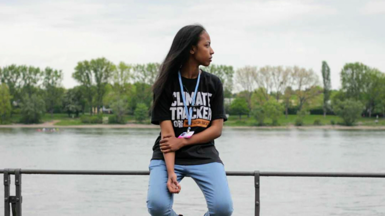 Eine junge Frau sitzt auf einem Geländer vor einem Fluss.