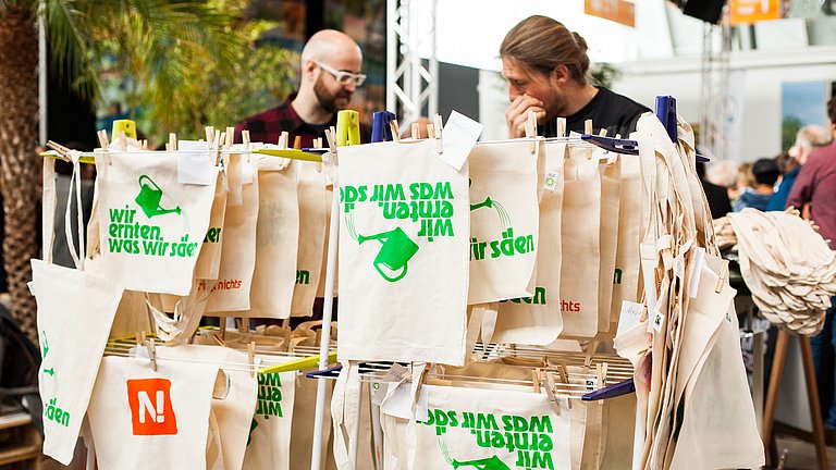 Siebdruckwerkstatt auf einer Messe: Jutetüten mit dem Aufdruck "Wir ernten was wir säen" sind aufgehängt zum Trocknen 