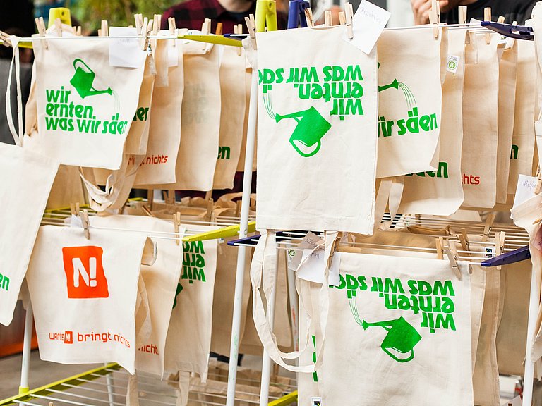 Siebdruckwerkstatt auf einer Messe: Jutetüten mit dem Aufdruck "Wir ernten was wir säen" sind aufgehängt zum Trocknen 