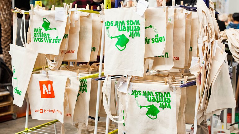 Siebdruckwerkstatt auf einer Messe: Jutetüten mit dem Aufdruck "Wir ernten was wir säen" sind aufgehängt zum Trocknen 