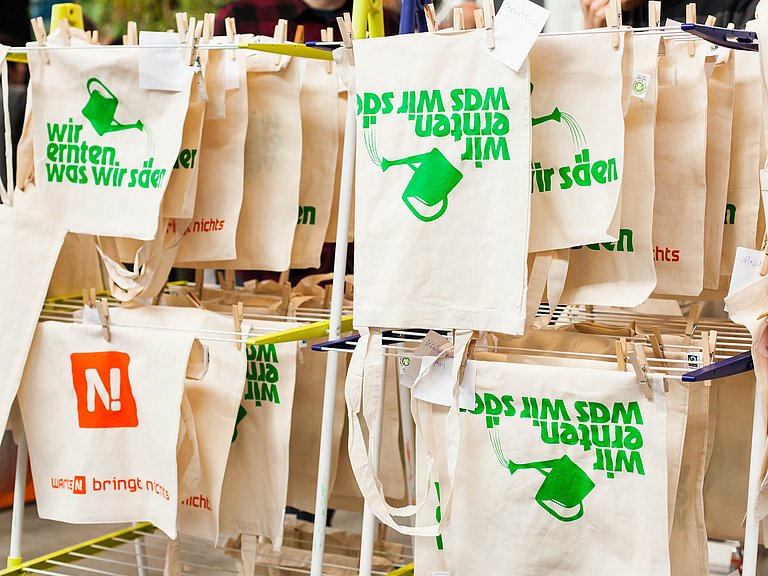 Siebdruckwerkstatt auf einer Messe: Jutetüten mit dem Aufdruck "Wir ernten was wir säen" sind aufgehängt zum Trocknen 