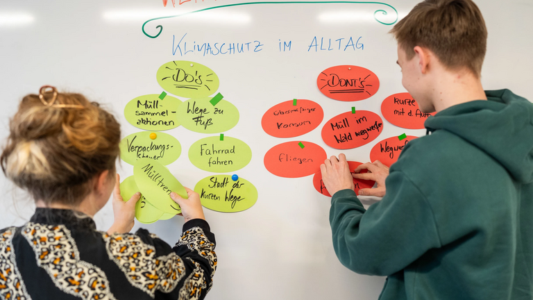 Zwei junge Personen pinnen Karteikarten an ein Whiteboard.