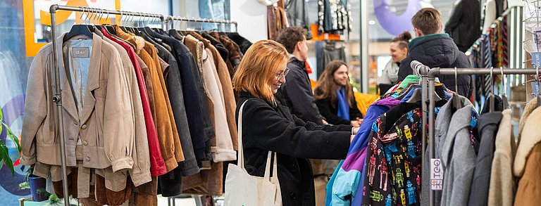 Junge Frau schaut sich Kleidung an. Im Hintergrund gibt es einen Workshop zum Thema Nachhaltiger Konsum.