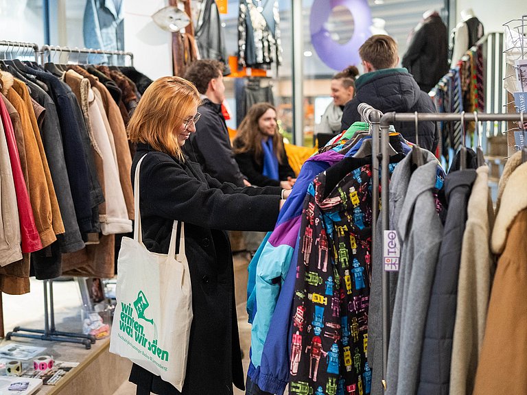 Junge Frau schaut sich Kleidung an. Im Hintergrund gibt es einen Workshop zum Thema Nachhaltiger Konsum.