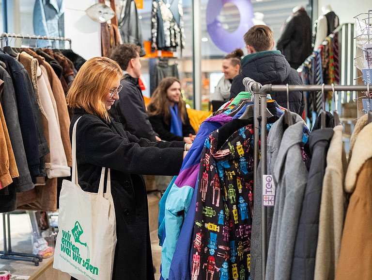 Junge Frau schaut sich Kleidung an. Im Hintergrund gibt es einen Workshop zum Thema Nachhaltiger Konsum.