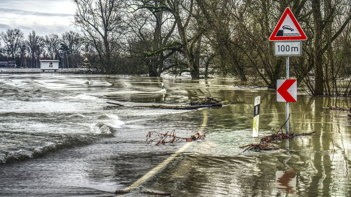 Überflutete Landstraße mit einem Verkehrsschild: Achtung in 100 Metern besteht für Autos Gefahr von der Straße abzukommen