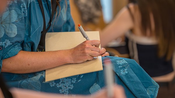Eine Frau schreibt in ein Notizbuch auf ihrem Schoß während einer Veranstaltung