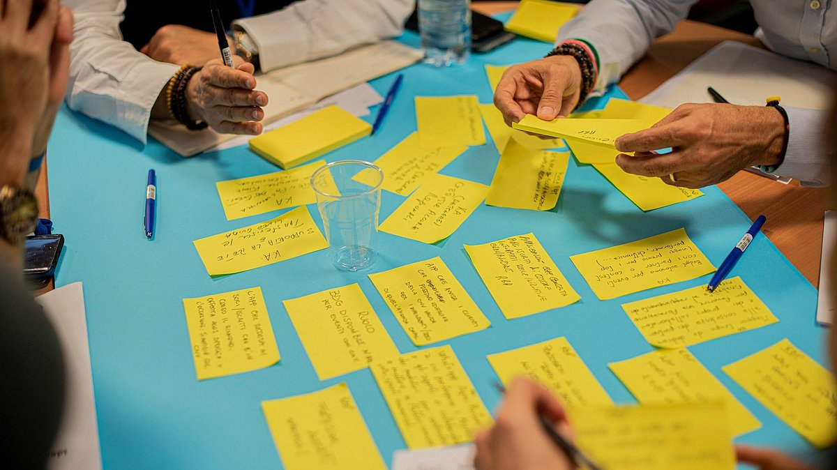 Mehrere Personen sitzen gemeinsam am Tisch und entwickeln Ideen, während sie gelbe Notizzettel und ein großes Plakat für ihr Team-Brainstorming nutzen.