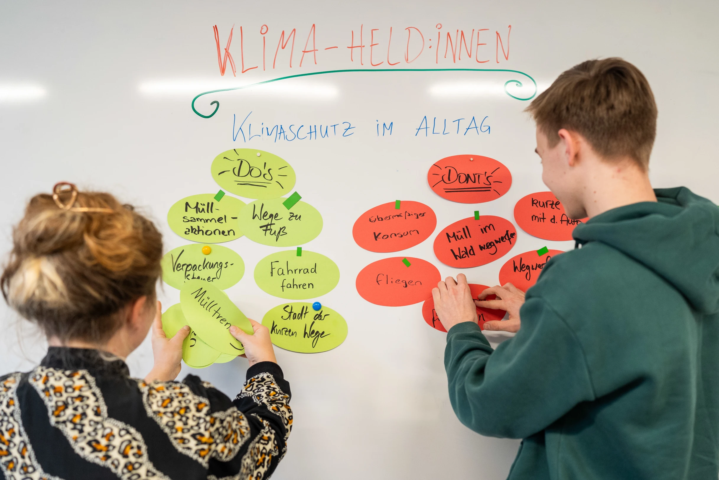 Zwei junge Personen pinnen Karteikarten an ein Whiteboard.